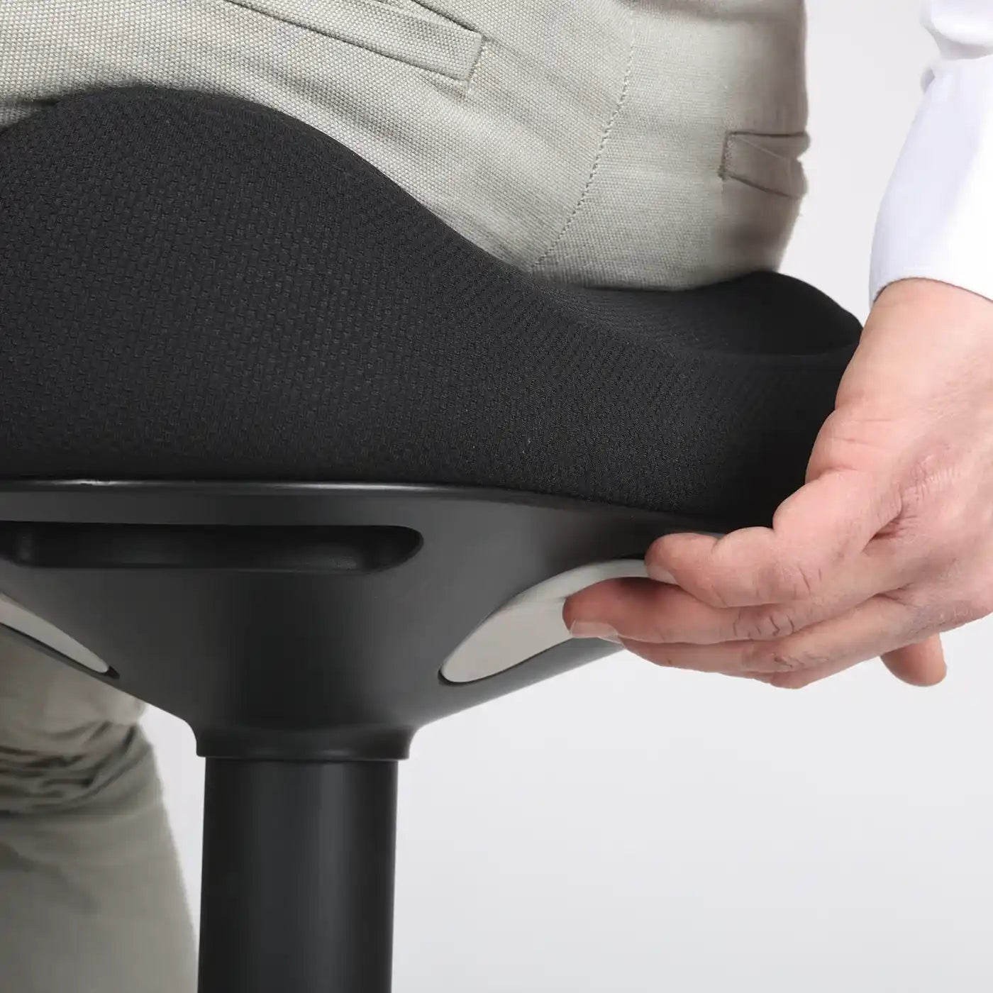 Close-up studio photo diagonal bottom view of a part of the seat of the Wobblez Soothe wobble stool in black and the knob visible with a person sitting on it with the hand on the knob with neutral white grey background