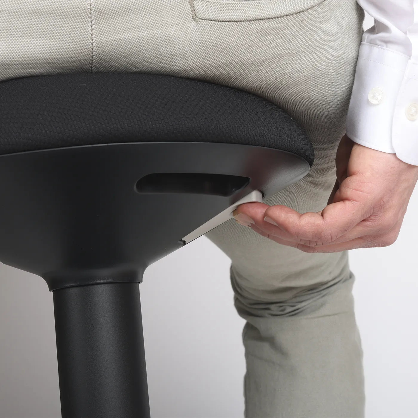 Close-up studio photo diagonal bottom view of a part of the seat of the Wobblez Savvy wobble stool in black and the knob visible with a person sitting on it with the hand on the knob with neutral white grey background