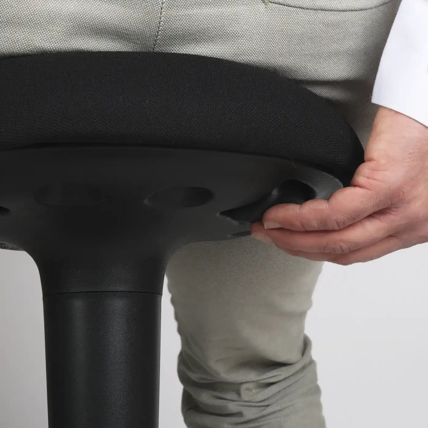 Close-up studio photo diagonal bottom view of a part of the seat of the Wobblez Spine wobble stool in black and the knob visible with a person sitting on it with the hand on the knob with neutral white grey background