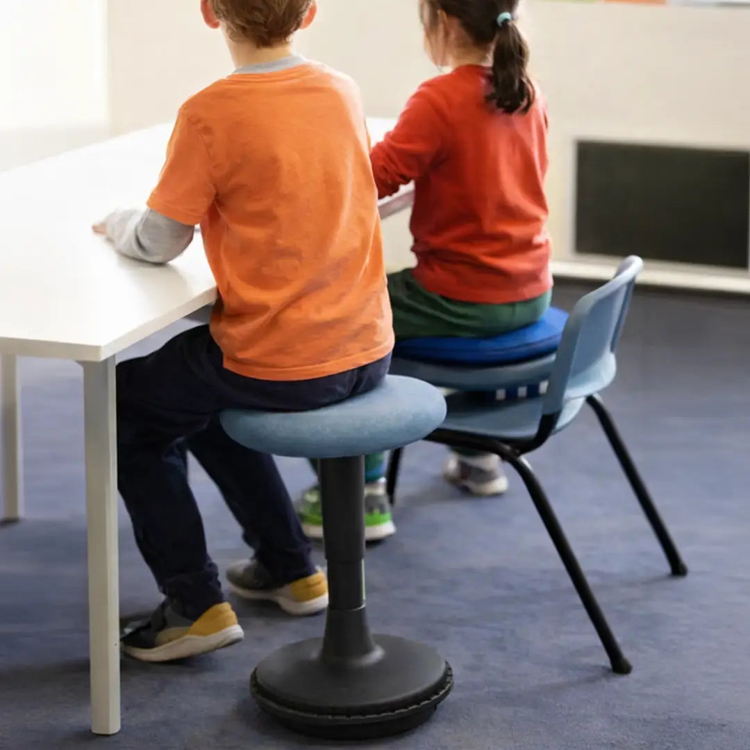 Two children at a table in a classroom setting. One kid on a wobble stool and the other on a regular chair with several cushions.