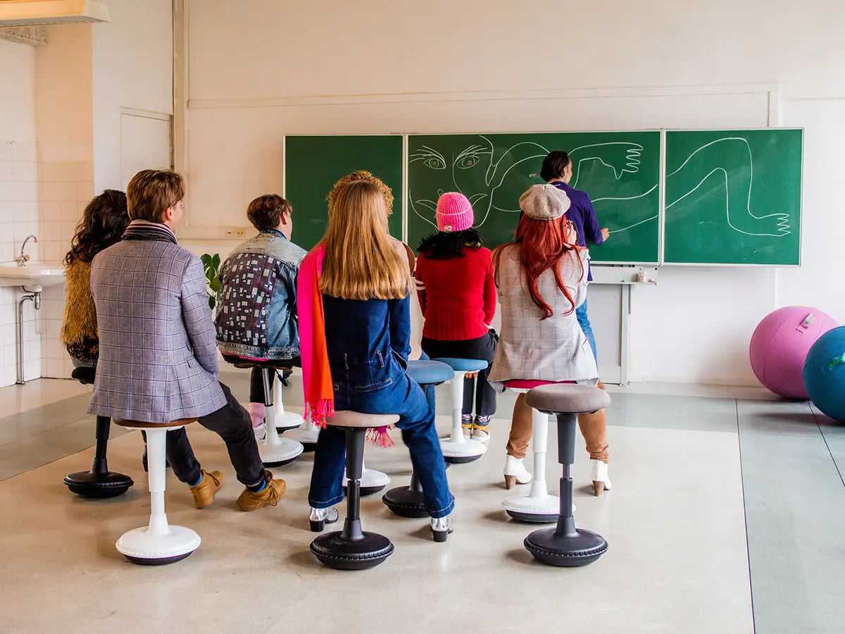 Group of students on Wobblez wobble stools in a classroom looking at a teacher in front of a blackboard with drawings.