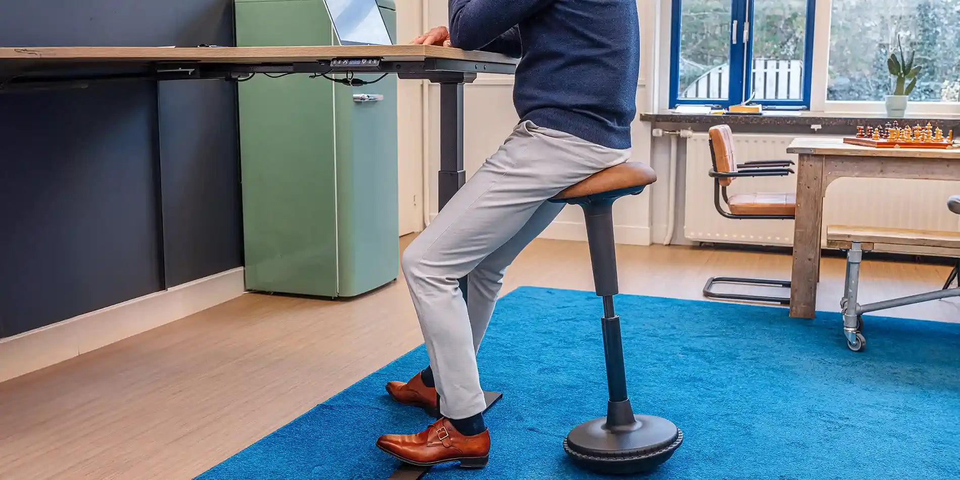 A man working on a standing desk in an office setting with a laptop and using a sit-stand Wobblez Spine wobble stool with a black frame and a faux leather cognac seat.
