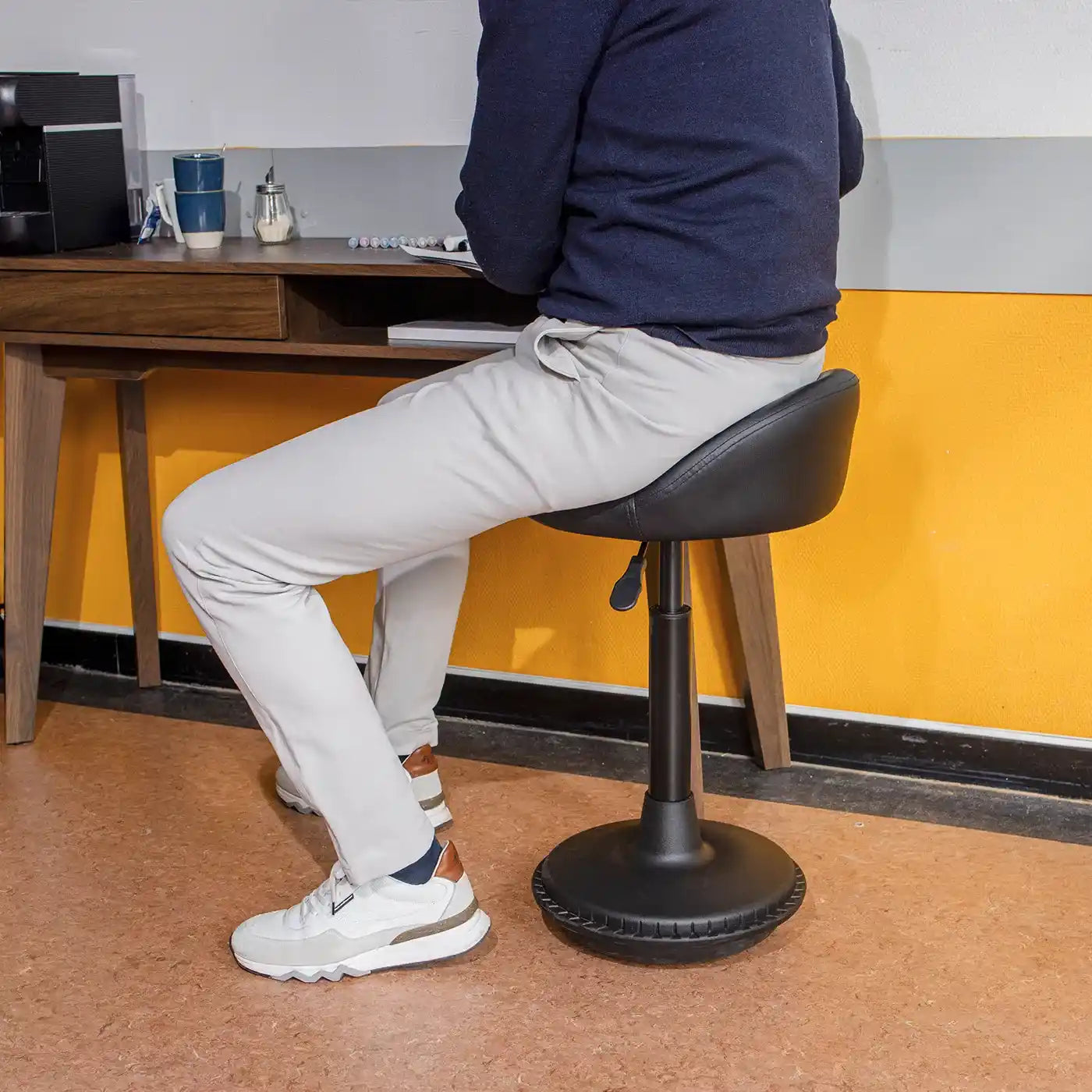 Person sitting on a black office Wobblez Settle wobble stool chair at a desk with a yellow wall in the background, wearing light pants and sneakers.