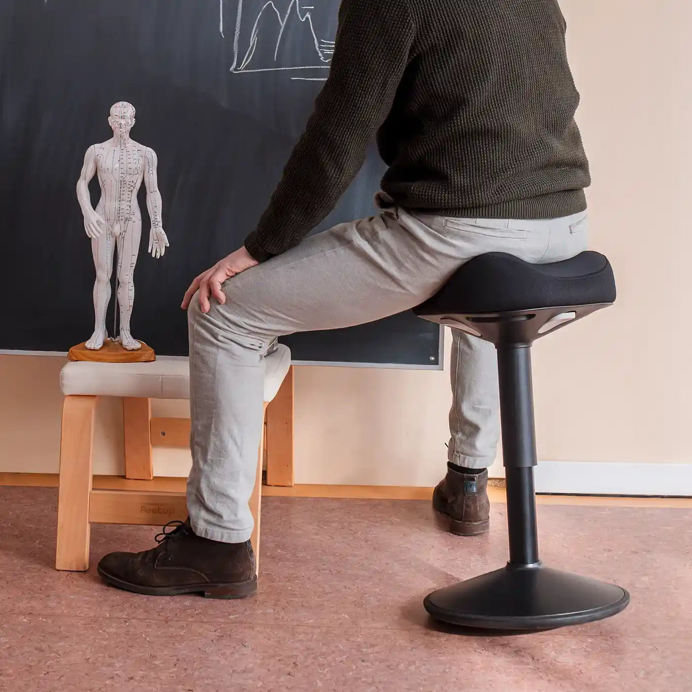 A teacher sitting on a black Wobblez Soothe wobble stool in a classroom writing on a chalkboard with an anotomic figurine