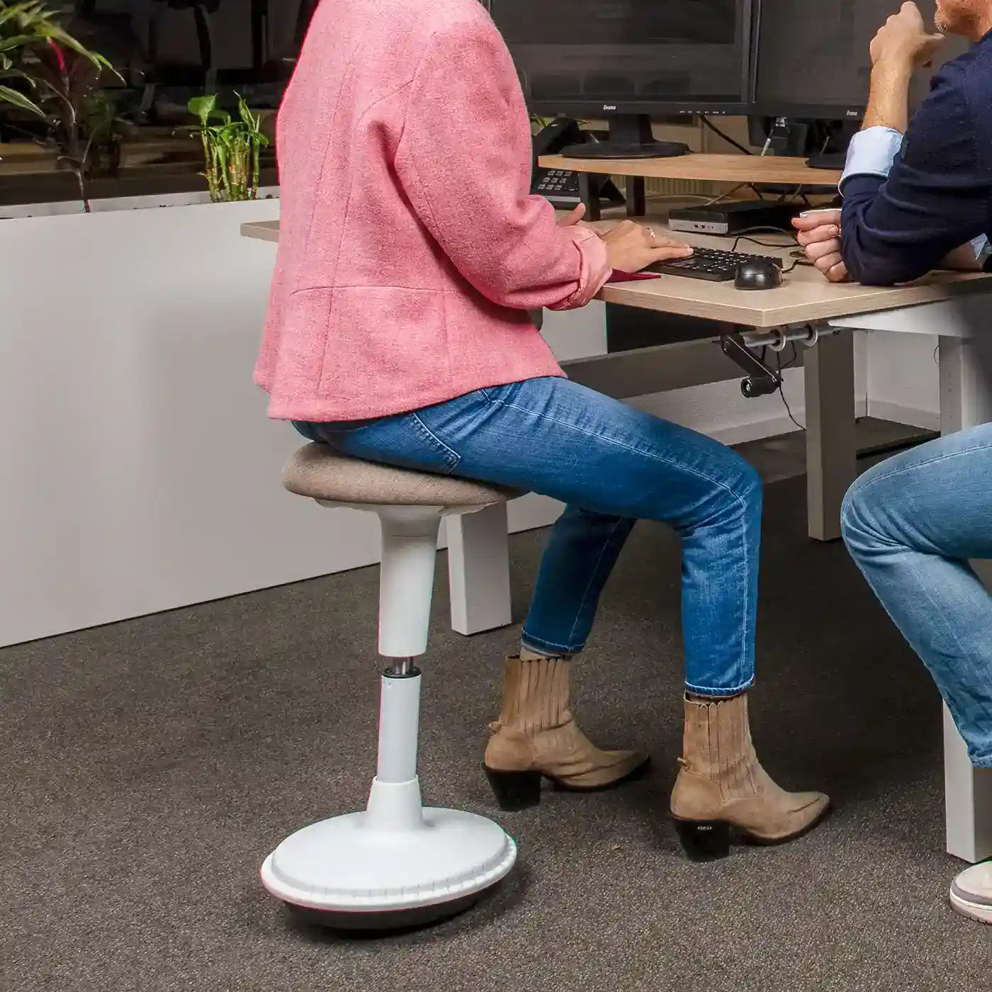 Person a pink jacket and blue jeans with brown boots sitting on a height-adjustable Wobblez Spine wobble stool with white frame and taupe seat at a working desk with another person.