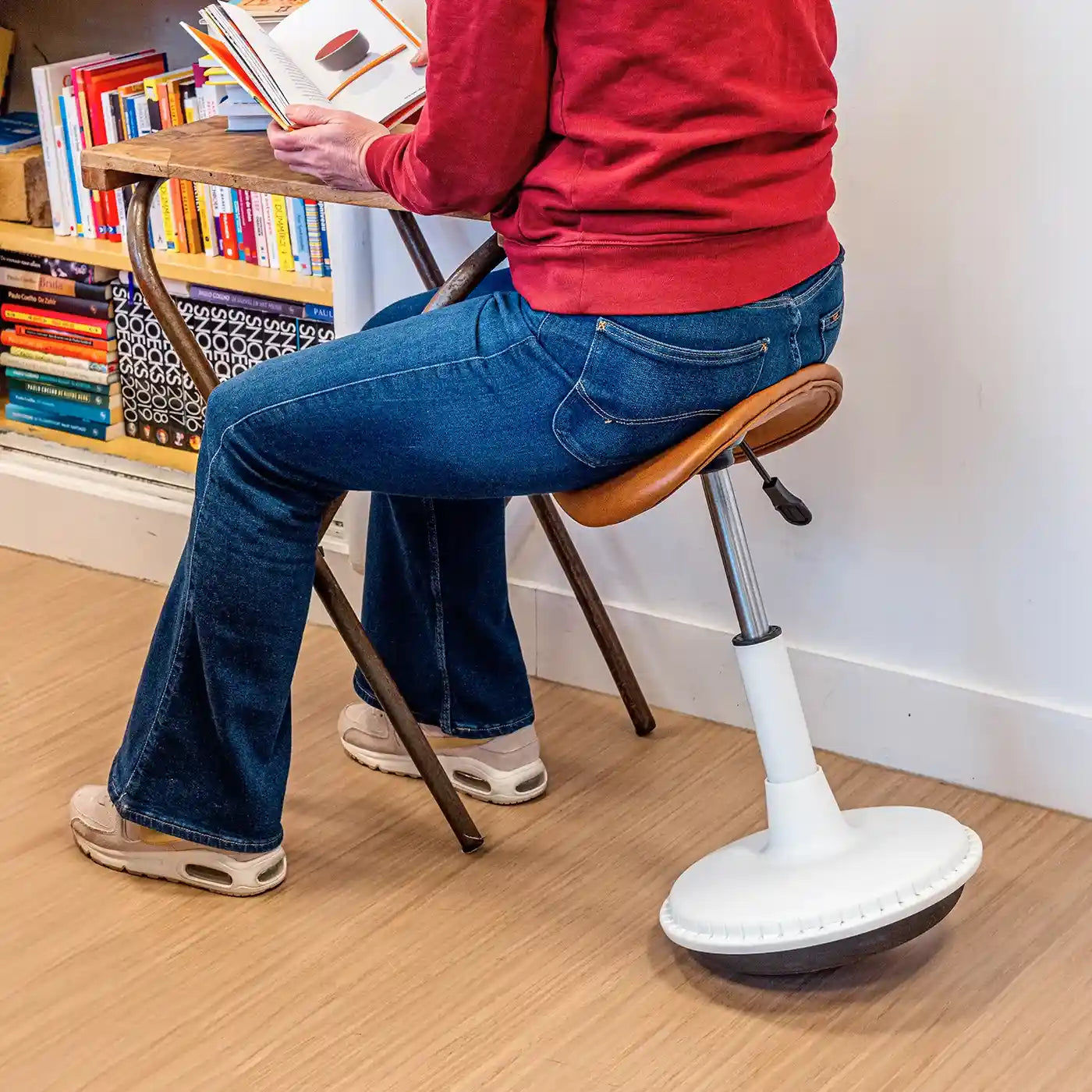 Style photography of person in red sweater and blue jeans using a Wobblez Stable wobble stool with faux cognac leather seat and a white frame reading a book on a wooden desk in a library