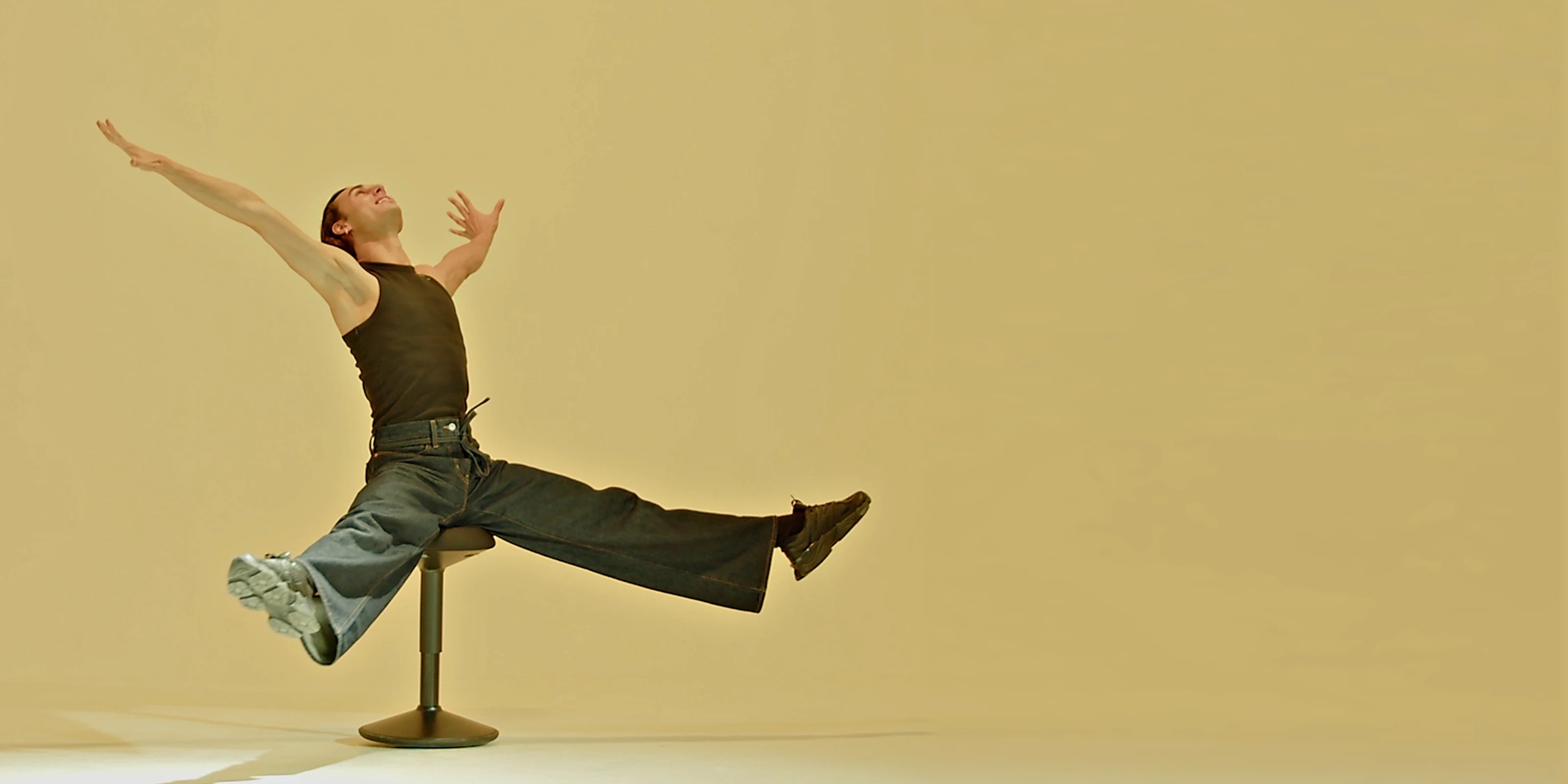 Wide view of person spreading arms and legs on a black Wobblez Savvy wobble stool set against a yellow gold background