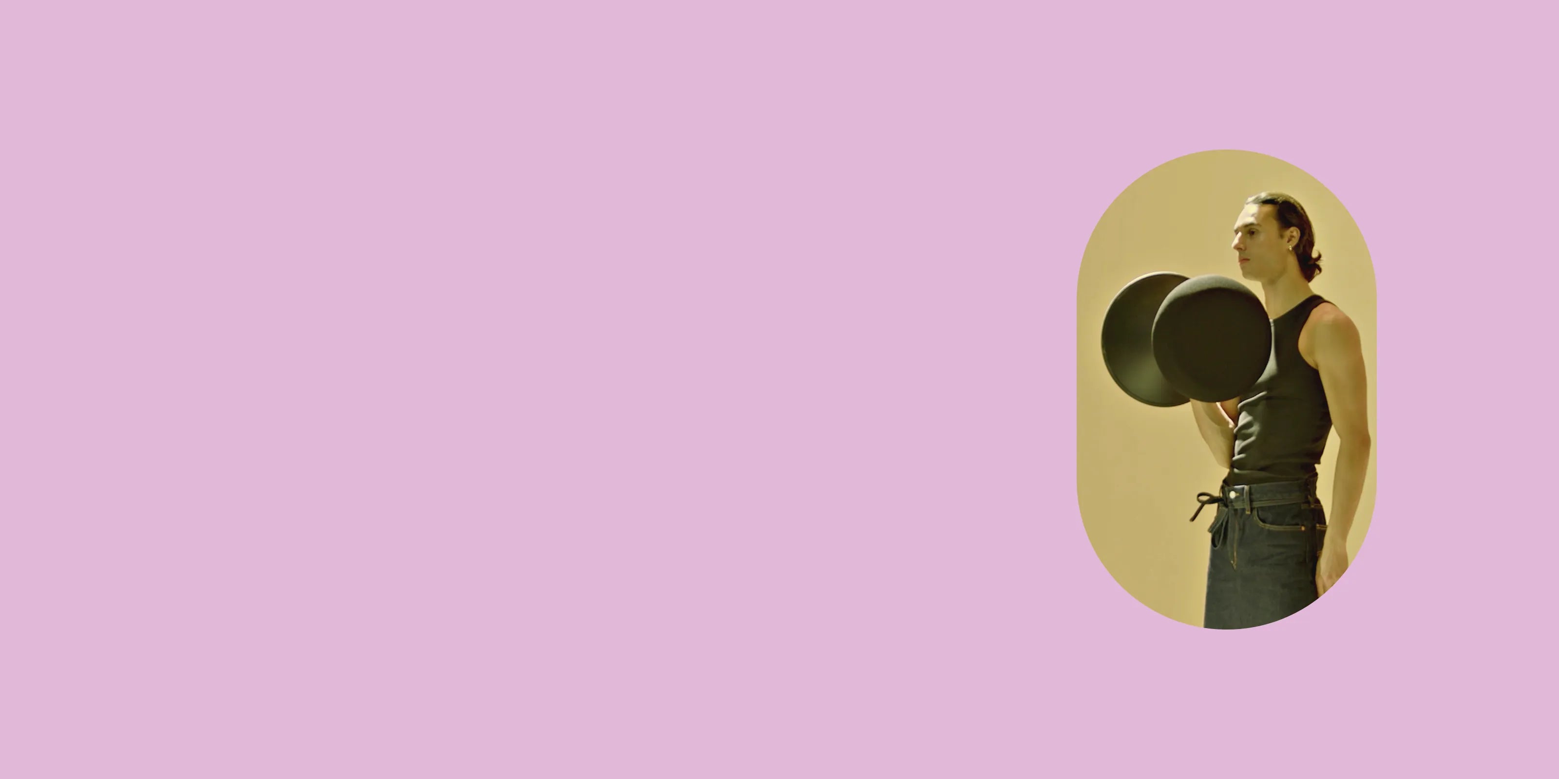A person in a black tank top and blue jeans lifting a black Wobblez Savvy wobble stool against a soft yellow background. The image is styled in a minimalist way with soft pink backgrouind emphasizing the subject and the objects.