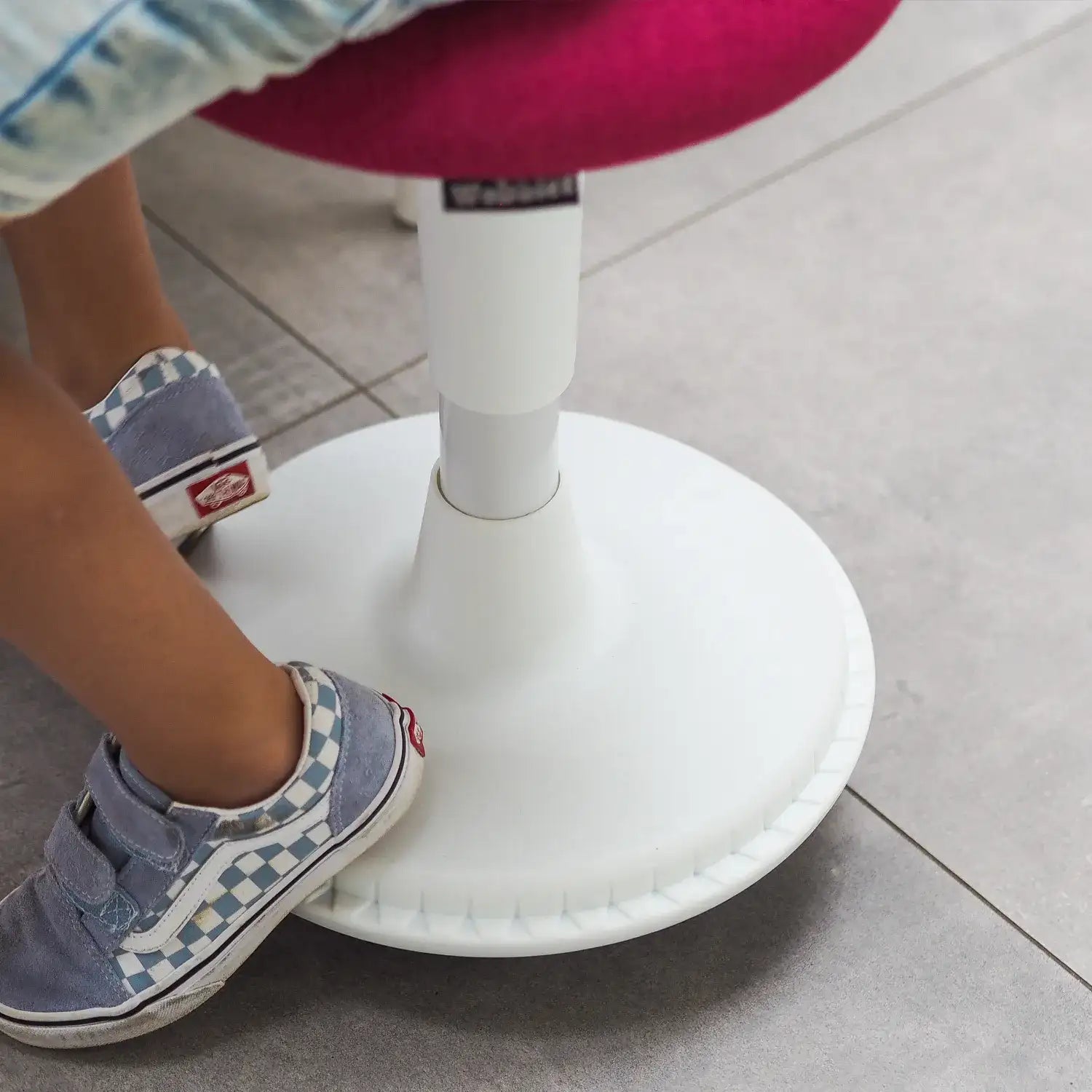 A Wobblez Spine wobble stool on a ceramic tiles floor with person on it, close-up birds eye view of the white frame with heavy wobble base and fuchsia fabric seat.