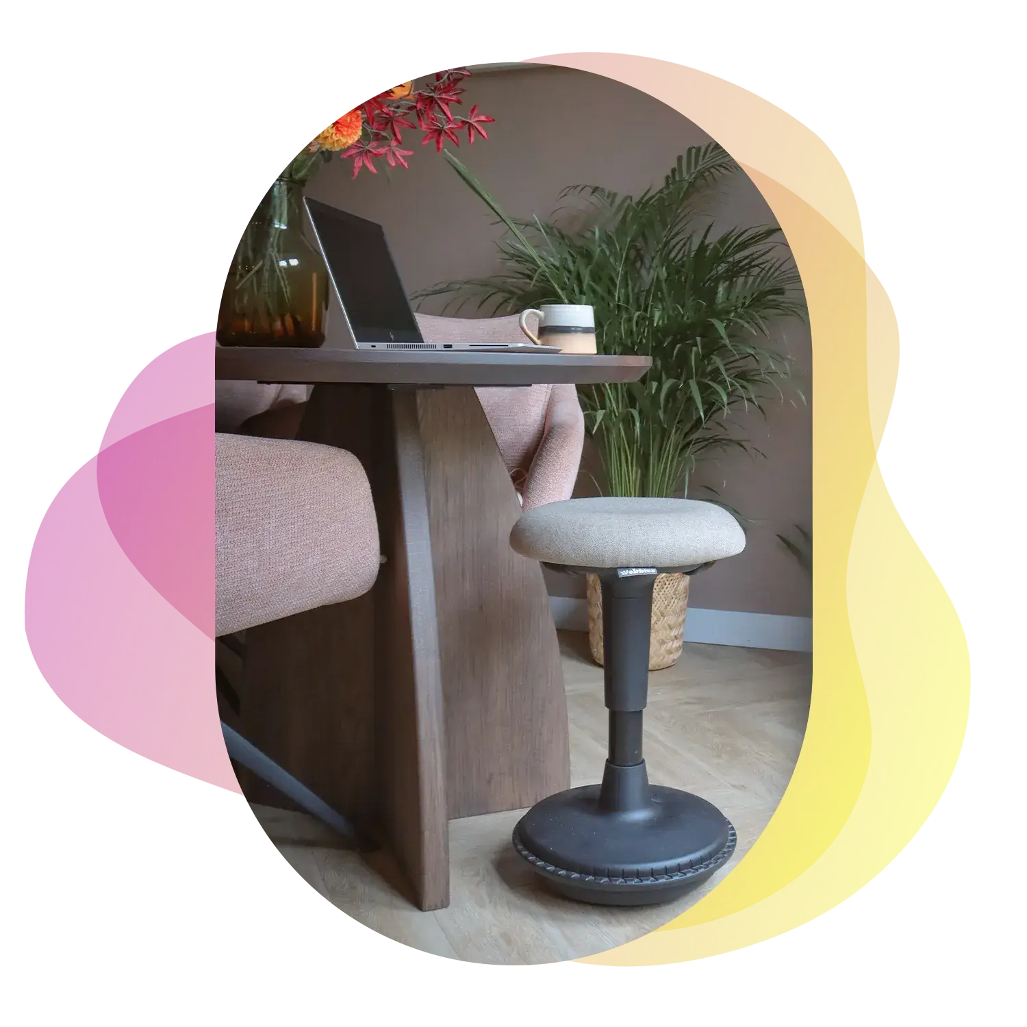 A Wobblez Spine black-pebble version positioned at a wooden dining table with a laptop and a coffee mug surrounded by Pink-black chairs and in living room with wooden floor and a plant against the wall and bright pink orange flowers on the table