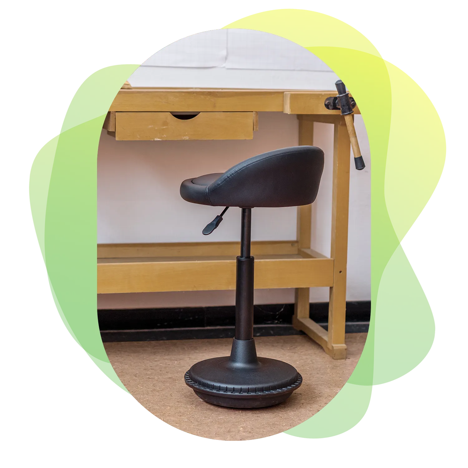 Image in the center of Wobblez graphic design styled blobs in green with a black Settle wobble stool in a workshop at a wooden worktable