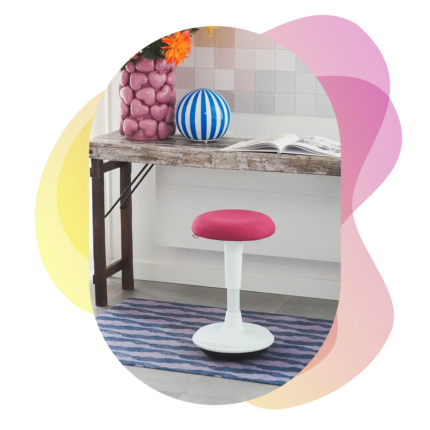 A white wobblestool with a pink fuchsia seat stands in front of a rustic wooden desk, decorated with a striped vase, a pink vase, and an open book, on a patterned rug.