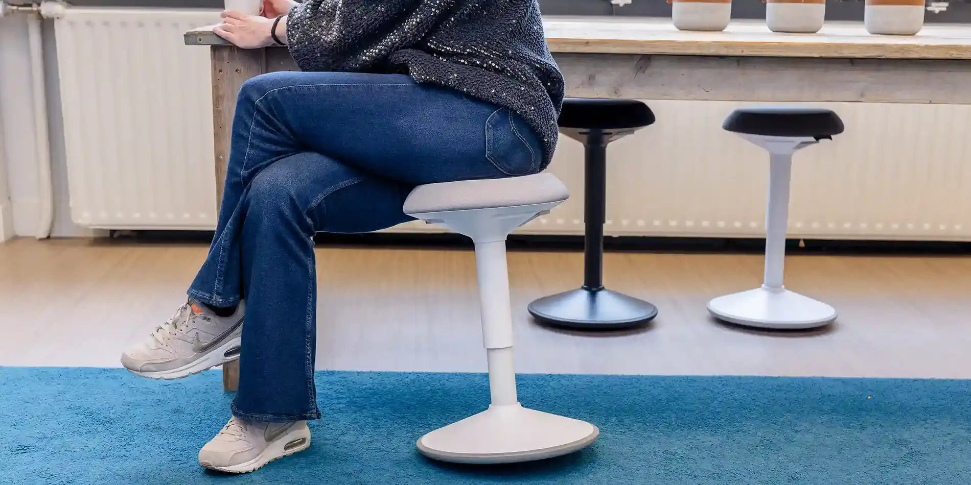 Person sitting on a height-adjustable white light gray Wobblez Savvy wobble stool in an office setting with an all black and white-dark gray variant on the other side of the wooden table